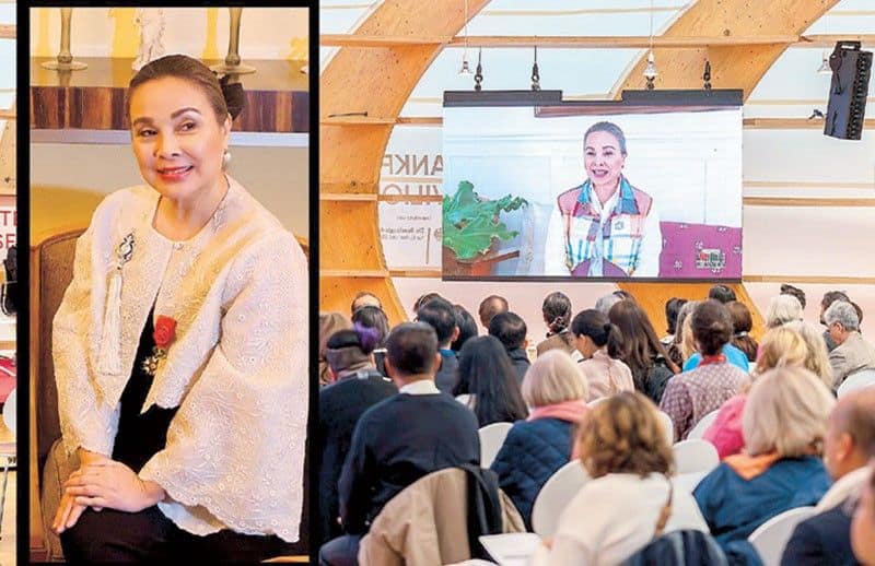 Sen. Legarda on a large screen wearing traditional attire. An audience is seated, watching attentively in a hall with wooden beams.