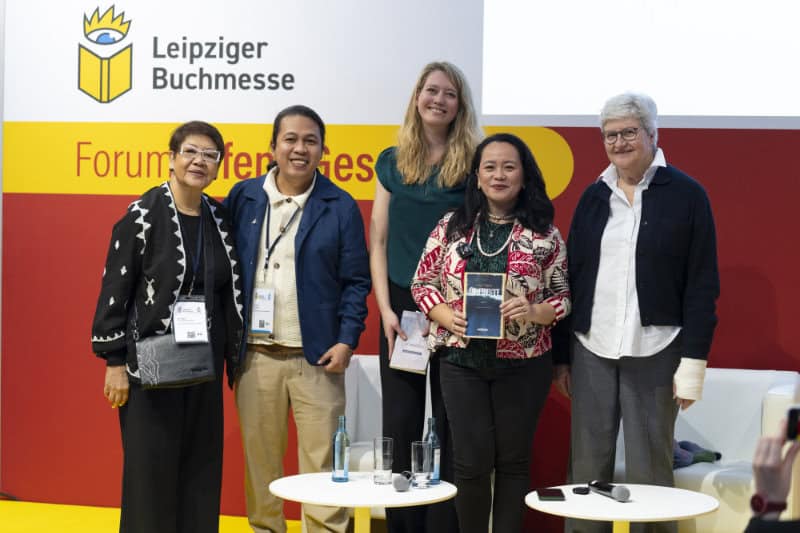 Five people stand together on stage at the Leipziger Buchmesse, posing for a photo. One person holds a book.