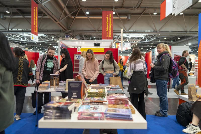 People browse books displayed on tables at the Leipzig Book Fair, with other attendees and exhibition booths visible in the background.