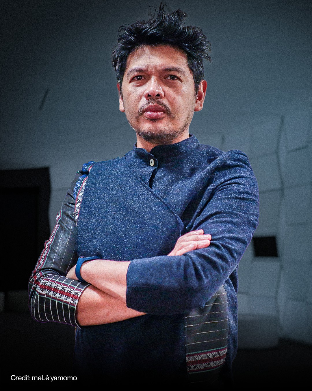 Artist meLê yamomo with dark hair and facial hair stands with arms crossed, wearing a navy jacket with patterned details.