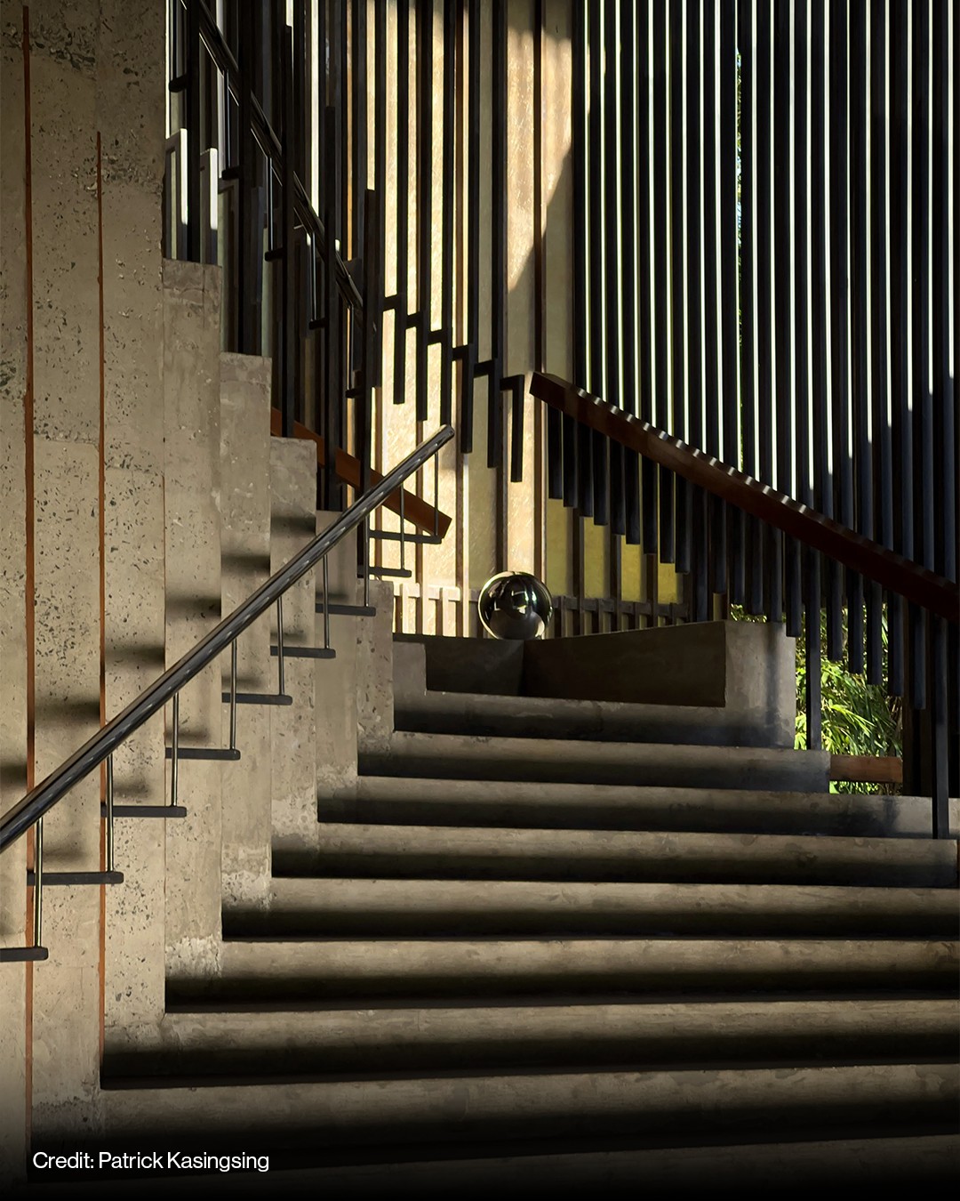 Concrete staircase with a metal handrail, vertical slatted wall, and sunlight casting shadows through the slats. A metallic sphere sits on a landing.