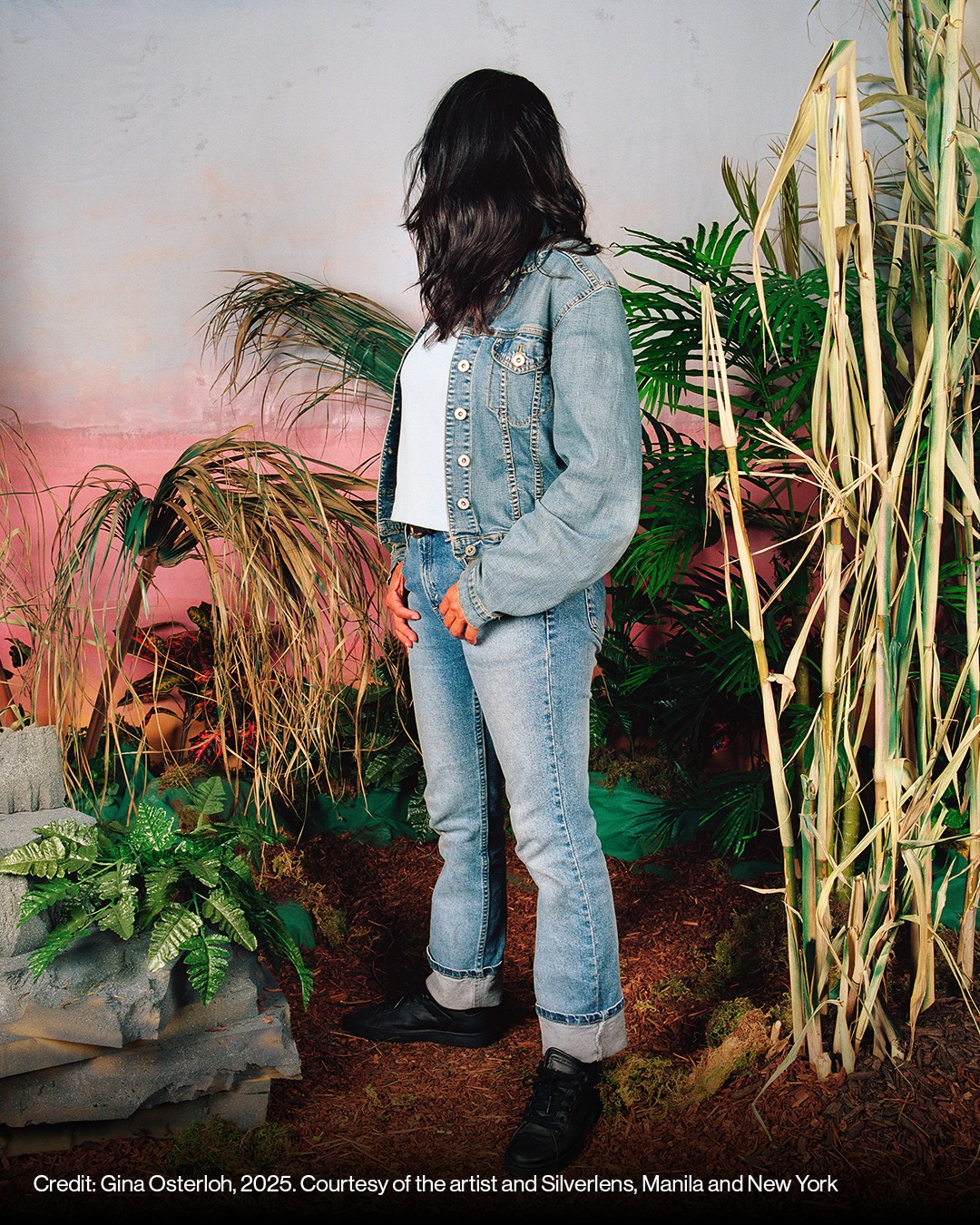 A person in a denim jacket and jeans stands with their head turned and hair obscuring their face, surrounded by plants and tall grasses in a staged indoor setting.