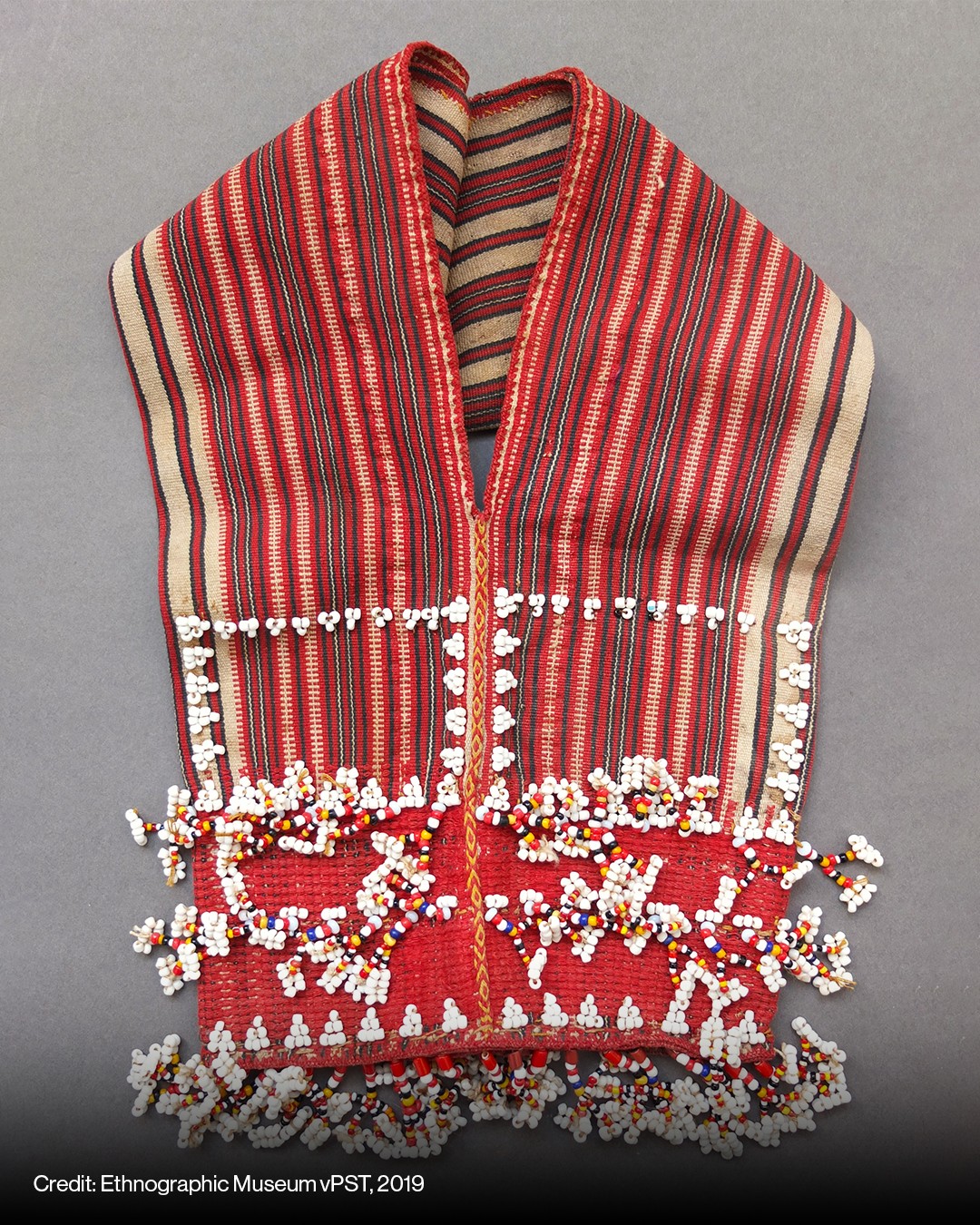 A traditional red woven textile with vertical stripes and geometric designs, decorated with clusters of colorful beads along the lower edge.