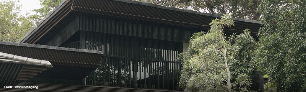 Modern building with large glass windows and black horizontal slats, partially obscured by green trees.