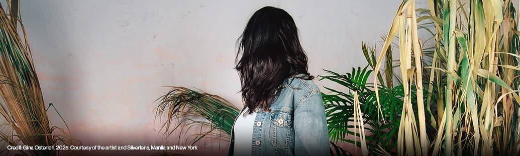 A person with long dark hair and a denim jacket stands facing sideways among tall plants against a plain wall.