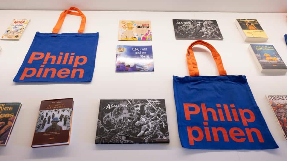 Two blue tote bags with "Philippinen" text are displayed among various books arranged on a white surface.